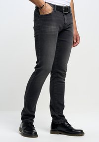 Jeans de mezclilla oscuros, de corte slim, con un ligero brillo. Presentan un diseño clásico de cinco bolsillos y un cinturón negro. Se combinan con zapatos negros de cordones.