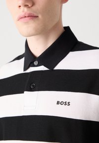 Svart og hvit stripete polo-skjorte med krage, med to knapper og en liten "BOSS" logo på venstre side. Bomullsstoff tekstur.