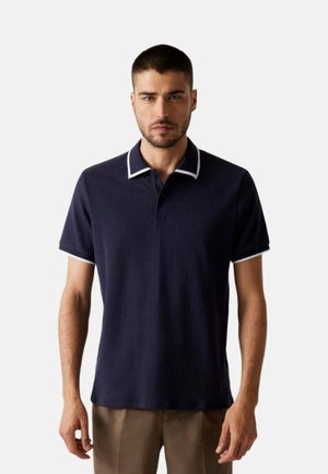 Uomo che indossa una polo a maniche corte color blu navy con rifiniture bianche sul colletto e sulle maniche, in piedi davanti a uno sfondo semplice.