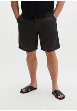 Man draagt zwarte shorts, zwart t-shirt en zwarte instapsandalen, staand met handen in de zakken op een witte achtergrond.