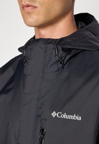Veste Columbia noire avec capuche, offrant une texture lisse et légère, des accents à cordon et une poche zippée à l'avant. Logo affiché de manière proéminente.