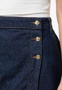 Dark denim skirt featuring gold spiral buttons along the side, with contrasting orange stitching and a smooth texture.