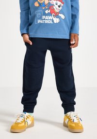Blaues Langarmhemd mit Paw Patrol Grafik, marineblaue Jogginghose mit gerippten Knieflexen und gelbe Sneaker mit weißen Akzenten.