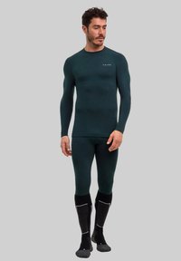 Zielony termiczny komplet bielizny bazowej z długimi rękawami i dopasowanymi legginsami, tkanina o fakturze, noszony z czarno-szarymi skarpetkami, minimalistyczny detal z logo.