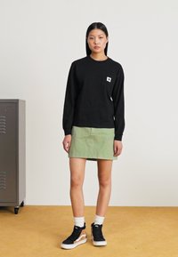 Schwarzes Langarmshirt mit einer Brusttasche, kombiniert mit einem grünen Denim-Rock. Das Outfit wird durch schwarze Sneaker mit weißen Akzenten ergänzt.