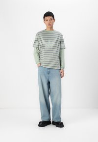 Camisa de rayas verdes y blancas de manga corta, superpuesta sobre una camiseta verde de manga larga, combinada con unos jeans de mezclilla sueltos de color azul claro y zapatos negros gruesos.