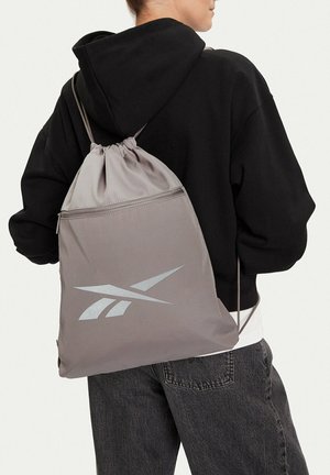 Persona con sudadera negra con capucha y mochila gris con cordón que tiene un gran logo blanco en la parte posterior, combinada con vaqueros gris oscuro.