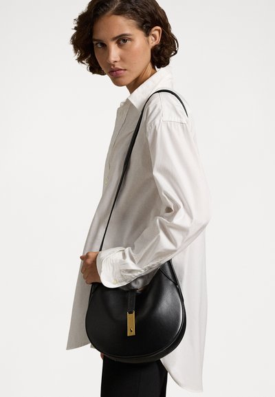 Bolso bandolera de cuero negro con forma redonda, correa ajustable y un acento de hardware dorado en el cierre.