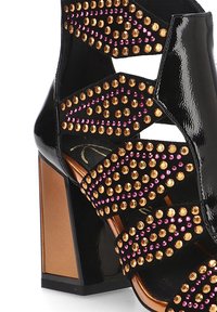 Sandalias de tacón alto de charol negro con acentos de tachuelas doradas y rosas, que presentan múltiples correas curvas y un diseño de tacón grueso.