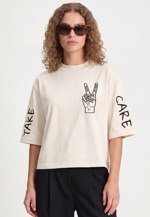Femme aux cheveux bouclés portant des lunettes de soleil écaille de tortue et une chemise beige avec un signe de paix et les mots "Take Care" sur les manches.