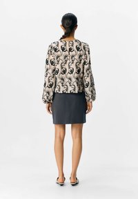 Femme avec des cheveux tressés portant un haut floral beige à manches longues, une jupe gris foncé et des sandales à talons noires et blanches, se tenant de dos.