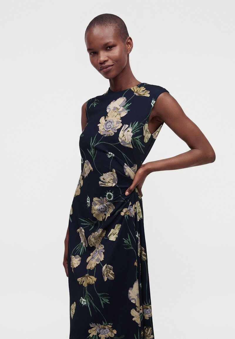 Woman standing with hand on hip, wearing a sleeveless navy dress with yellow floral print against a plain light background.