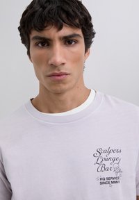Joven con cabello corto y rizado oscuro, vistiendo una camisa blanca con el texto "Scalpers Lounge Bar HQ Servicio Desde MMVII" en el pecho.