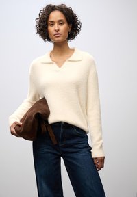 Maglione soffice di colore crema con scollatura a V e colletto, abbinato a jeans denim blu scuro. Tiene in mano una borsa in suede marrone.
