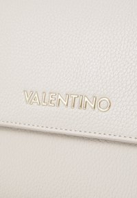 Hvit teksturert lærveske med et gull "VALENTINO"-logo. Overflaten viser fin kornstruktur og pent broderi langs kantene.
