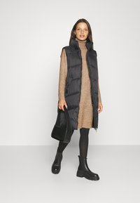 Svart vadderad väst med hög krage, bärs över en brun stickad klänning, i kombination med svarta leggings och kraftiga svarta ankelstövlar. Svart axelväska.