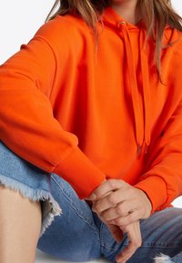 Pomarańczowy bawełniany hoodie z kapturem ściąganym sznurkiem, elastycznymi mankietami i luźnym krojem. Noszony z jasnoniebieskimi wystrzępionymi dżinsowymi szortami.