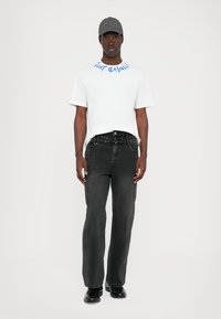Hvit t-skjorte med korte ermer og blå trykk. Svarte jeans, rett kutt, med frontlommer. Svarte sko og grå caps fullfører antrekket.