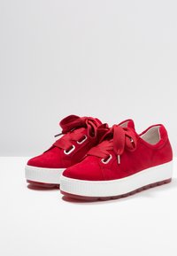 Baskets en daim rouge avec une semelle en caoutchouc blanche, dotées de larges lacets en tissu, d'œillets métalliques et d'une semelle texturée et surélevée pour une meilleure adhérence.