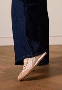 Sneaker in suede e pelle rosa chiaro con accentuazioni bianche e suola in gomma testurizzata, indossata con pantaloni di jeans scuri, parzialmente visibili.