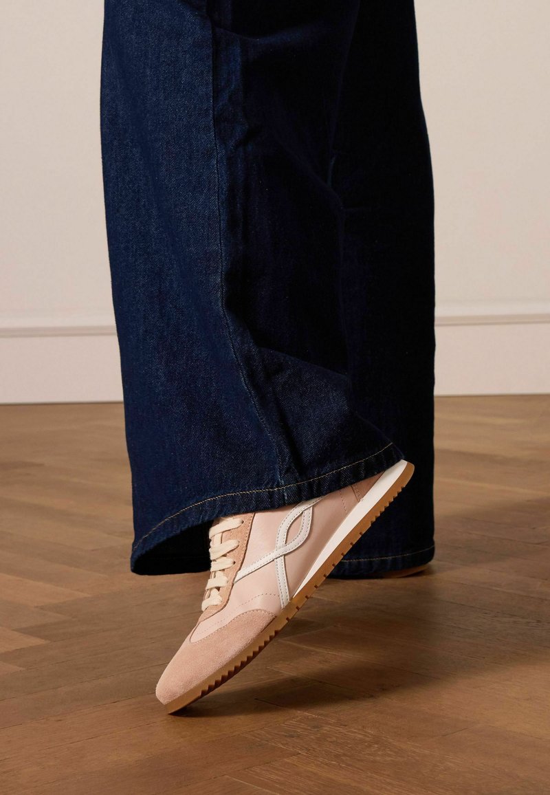 Sneaker in suede e pelle rosa chiaro con accentuazioni bianche e suola in gomma testurizzata, indossata con pantaloni di jeans scuri, parzialmente visibili.