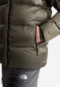 Person wearing olive green puffer jacket with zipped pocket and grey The North Face pants, hand inside jacket pocket.