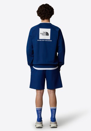 Marineblaues Sweatshirt mit weißem Logo auf dem Rücken, kombiniert mit passenden Shorts. Akzentuiert durch blaue Socken mit Text und grauen Turnschuhen.