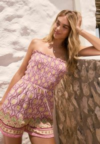 Trägerloser pinker Jumpsuit mit gerafftem Oberteil, gelbem und grünem Blumenmuster sowie strukturierten Saumdetails. An einer Steinwand getragen.