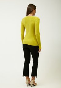 Maglione a maniche lunghe a coste di un vivace giallo, abbinato a pantaloni neri con pizzo lungo l'orlo e scarpe con tacco nude.