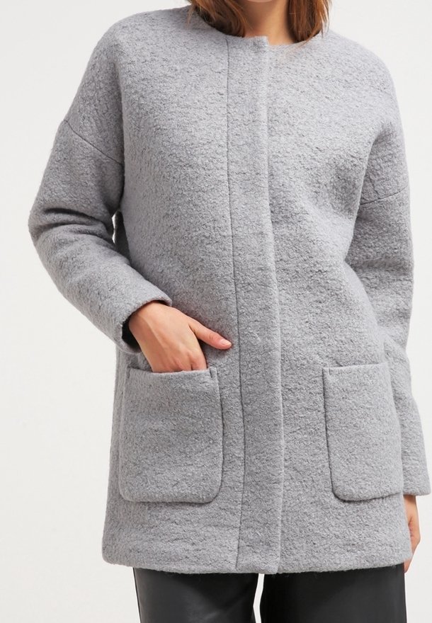 Femme portant un manteau gris clair texturé avec deux poches avant, une main dans une poche, associé à un pantalon noir.