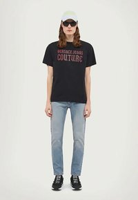 Person som har på seg en svart "Versace Jeans Couture" t-skjorte, lyseblå jeans, svarte joggesko med hvite såler, svarte solbriller og hvit caps.