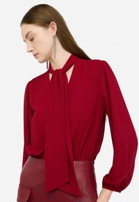 Blusa rossa con un colletto a cravatta, maniche lunghe a sbuffo e vita arricciata. Il tessuto appare liscio, abbinato a una gonna in pelle bordeaux.