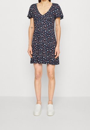 Kvinde iført en mørkeblå kortærmet kjole med små hvide og røde blomsterprint og hvide slip-on sko.