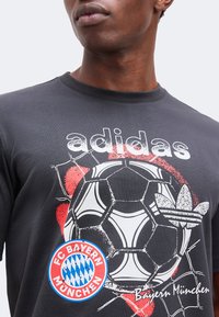 Κοντινό πλάνο ενός ατόμου που φοράει μαύρο T-shirt Adidas με μπάλα ποδοσφαίρου, λογότυπο FC Bayern München και κόκκινα γραφικά στοιχεία.