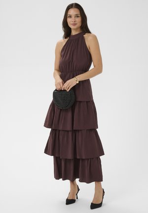 Vrouw draagt een mouwloze bruine maxi-jurk met lagen, houdt een zwarte gevlochten clutch vast en draagt zwarte hakken en een gouden armband tegen een effen achtergrond.