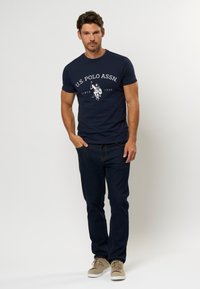 Marineblå t-skjorte med hvit "U.S. Polo Assn." logo, korte ermer, kombinert med mørkeblå jeans og beige sneakers. Enkel, uformell design.