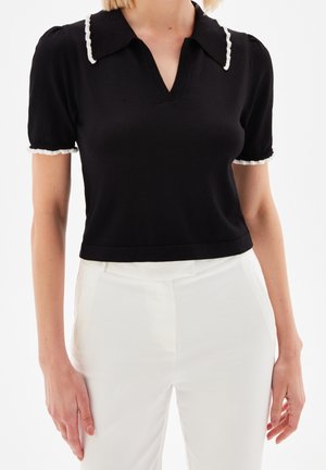 Černý top s krátkým rukávem a výstřihem do V, s kontrastním bílým ribbed límcem a manžetami, spárovaný s bílými přizpůsobenými kalhotami. Hladká textura.