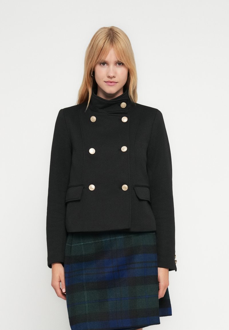 Μαύρο διπλόσπαστο σακάκι με χρυσά κουμπιά ως διακοσμητικά στοιχεία, με ψηλό γιακά και δύο μπροστινές τσέπες, φορεμένο πάνω από μια πράσινη και μπλε καρώ φούστα.