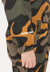 Kamerasuit i grønt og brunt med elastiske manchetter, en lynlåslomme og "W-PRO 10000" branding på ærmet.
