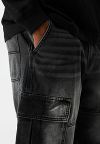 Svarta denim cargobyxor med sidofickor, en texturerad finish och synliga sömsdetaljer. Närbild som fokuserar på material och design.