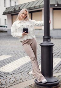 Bílá džínová bunda, béžové úzké kalhoty a bílé tenisky. Stojí vedle černého lampového sloupu na dlážděné cestě a drží telefon.