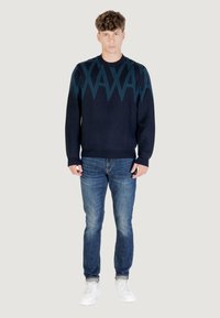 Jeune homme debout, portant un pull marine avec un motif géométrique bleu, un jean bleu et des baskets blanches sur un fond uni.