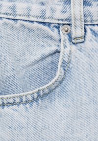 Poche de jean en denim bleu clair avec rivet métallique et passant de ceinture, montrant la texture détaillée du tissu et les coutures.