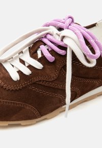 Rjav semiš športni čevelj z belimi in vijoličnimi vezalkami, zvezanimi v ohlapen lok, na svetli podplati na belem ozadju.