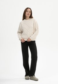 Cremfarbener Strickpullover mit Rautenmuster; lockere Passform. Kombiniert mit schwarzer gerader Hose und hellgrauen Slippern.