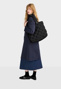Borsa tote nera trapuntata con texture imbottita e doppie tracolle, portata da una persona con un cappotto blu navy e una gonna di jeans su uno sfondo semplice.