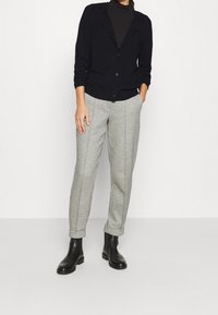 Cárdigan negro con botones, superpuesto a un suéter de cuello alto negro. Pantalones de cuadros grises con un corte entallado y puños, combinados con botas negras hasta el tobillo.