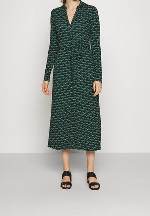 Vrouw draagt een midi-jurk met lange mouwen, knopen, zwart-groen patroon, met een ceintuur ter hoogte van de taille en zwarte sandalen met open teen.