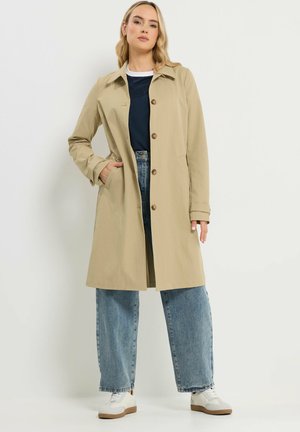 Femme portant un long manteau beige boutonné, une chemise bleu marine foncé, un jean bleu à jambes larges et des baskets blanches, debout avec une main dans la poche.