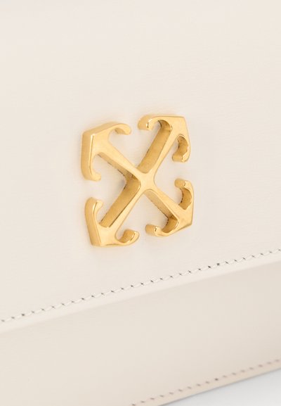 Έμβλημα από χρυσό μέταλλο με τέσσερα βέλη που ενώνονται στο κέντρο σε λείο, λευκό δερμάτινο φινίρισμα με ραφές.